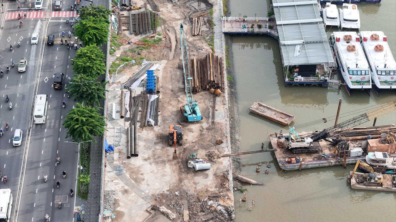 Aerial shot of a construction site by a river with adjacent road traffic and docked boats.